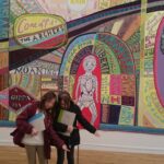 2 young girls bowing in front of UK mural with caricature of Queen